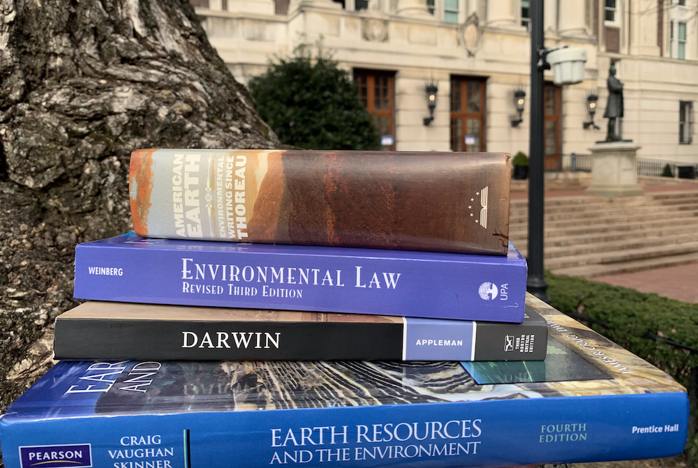 image of environmental science and law books at the foot of a tree in front of a building on a university campus. 